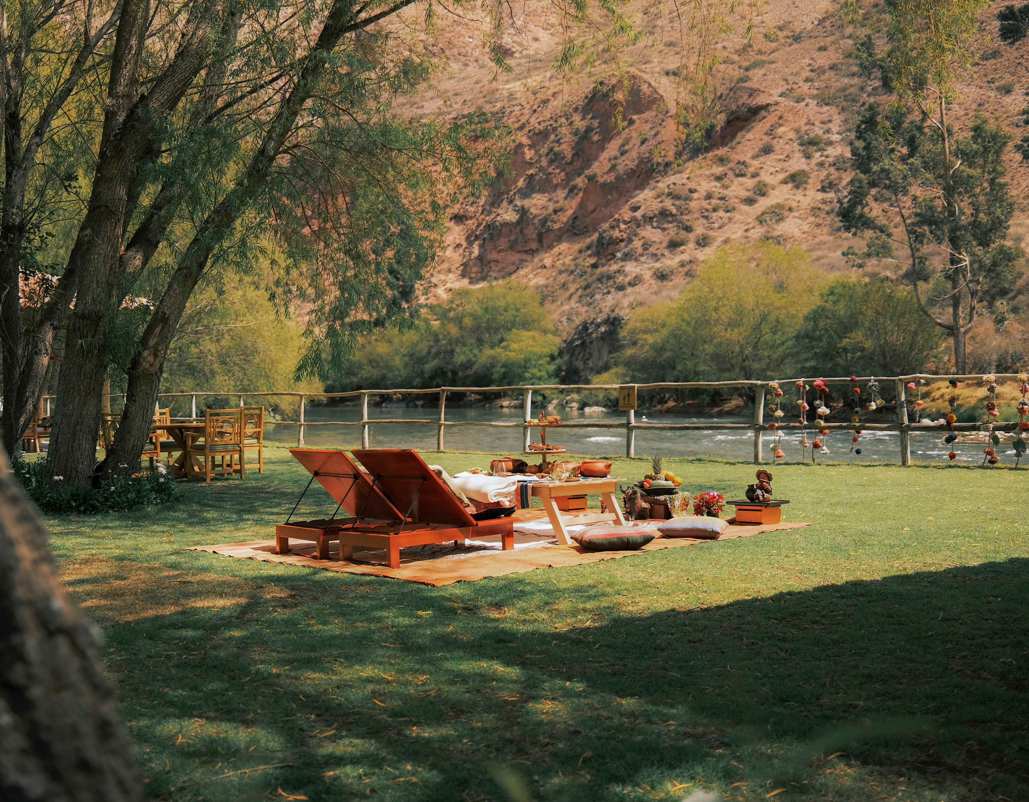 Two wooden loungers recline in front of a picnic table laden with food on a blanket with cushions, set on lawn facing the river.