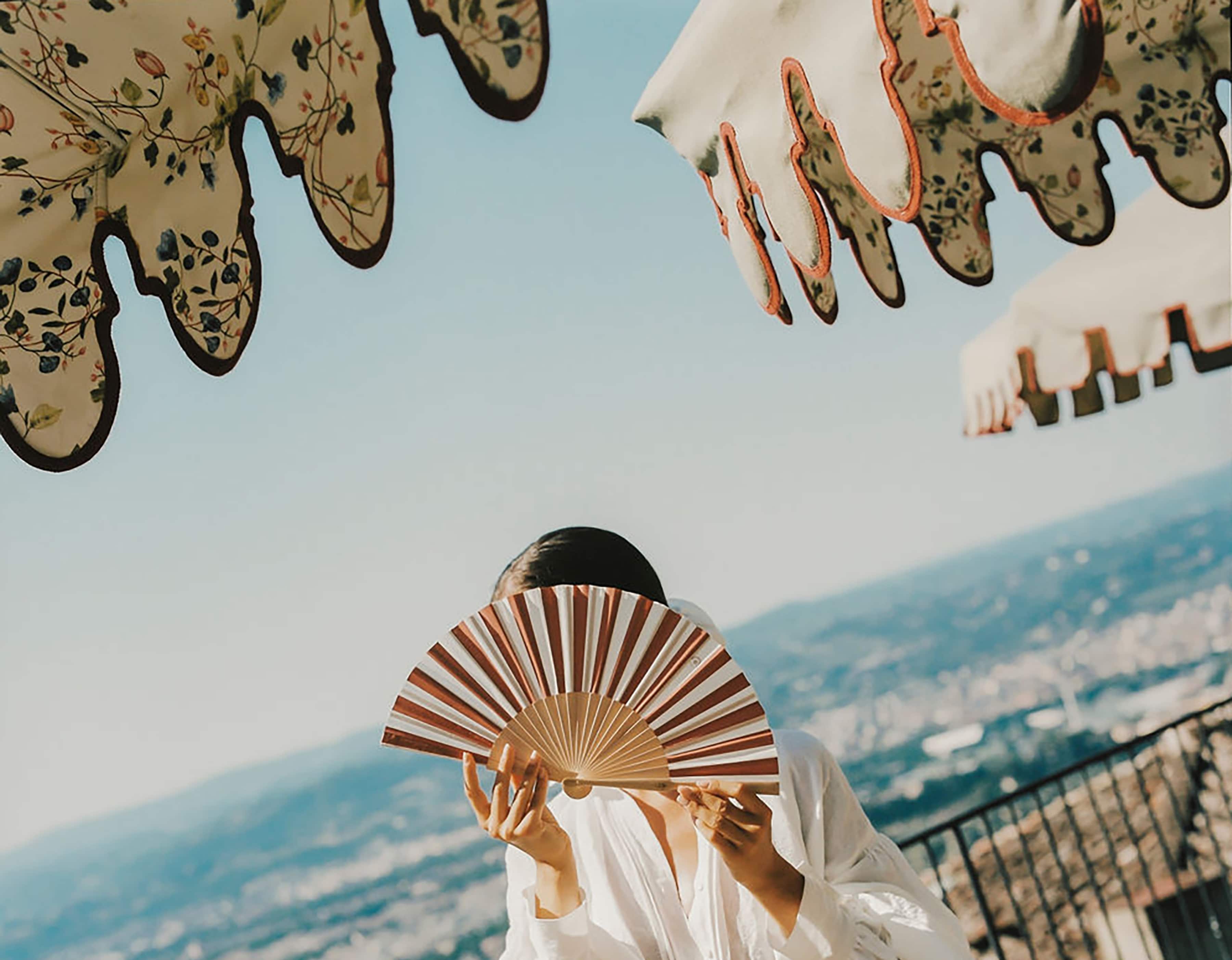 A woman in a diaphanous short white dress holds a fan in front her face as she stands in sunny gardens between parasols.