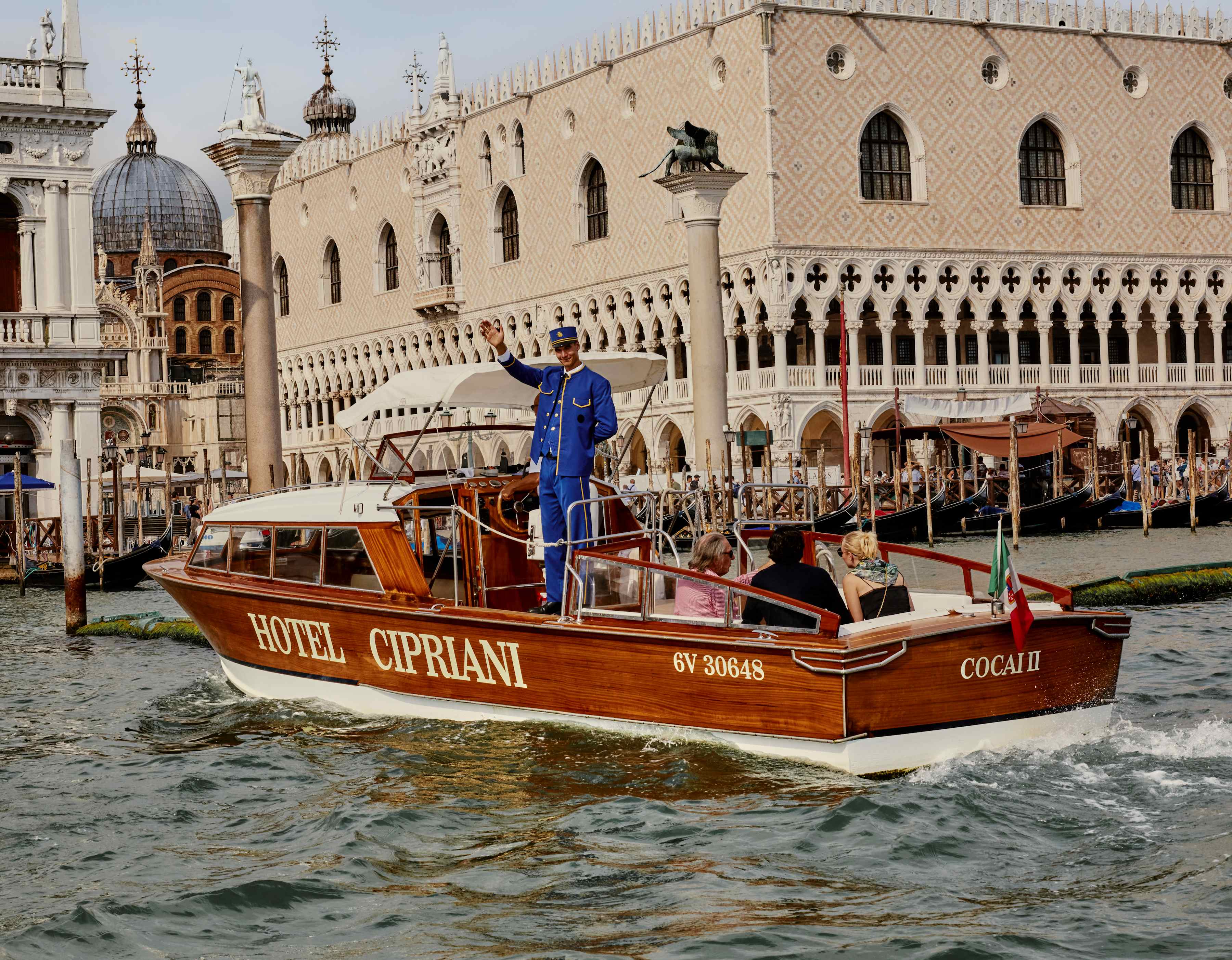 A man in a blue uniform waves as he stands on Hotel Cipriani's wooden boat with three guests, docking by the Doge's Palace.