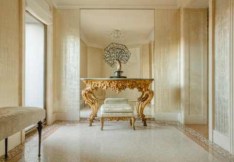 An ornamental tree-shaped lamp rests atop an ornately-carved rococo gilt dressing table with stool in front of a wall mirror.