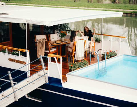 Guests relax round a table on the deck while the boat is moored, with a gangplank to the canal bank.