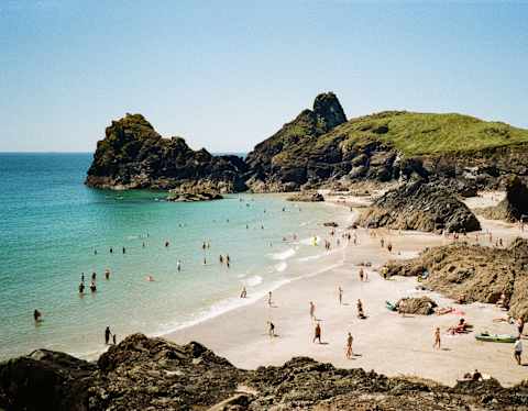 People relax and swim at a sandy beach surrounded by rocky cliffs and clear blue water under a sunny sky. Green hills rise in the background.