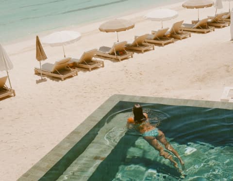 High angle shot in soft hues of a woman floating in the emerald water of Uchu's infinity pool overlooking the beach and sea.