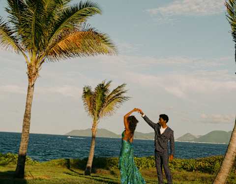 A man in a suit twirls a woman in a satin green dress as they dance between palm trees on lawn overlooking the sea.