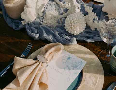 Close-up of a cream napkin on a capiz placemat at a wedding table with coral centrepieces arranged on wave-like blue cloth.