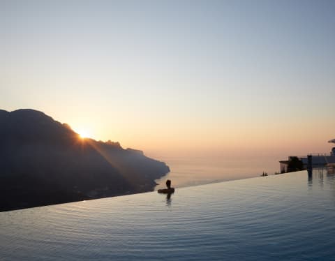 The last cathedral rays of sunshine dip below the Lattari Mountains as a woman gazes on from an infinity pool’s edge
