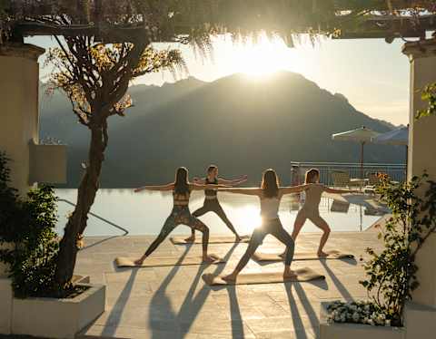 A group of four women adopt the Warrior Pose on mats by the pool as the sun rises behind Amalfi's coastal hills.