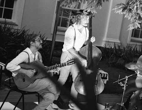 Two musicians perform outdoors; one sits playing guitar while the other stands playing a double bass. Both wear light shirts and braces. Sunlight and shadow fall across the scene. A building with windows is visible in the background.