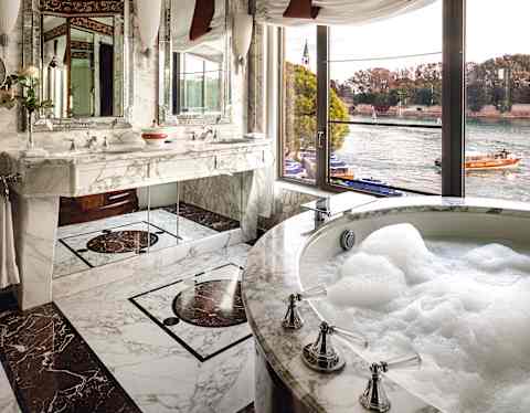 Soap bubbles fill the circular bath of a tub and russet marble bathroom. Outside large windows, the Hotel launch sets sail