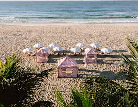Three red-striped cabanas and ten pairs of loungers shaded by white parasols face the sea in a view of the beach over palms.