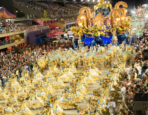 High-angle view of the Sambadrome Parade with dancers in straw-like costumes behind a blue and gold float, flanked by crowds.