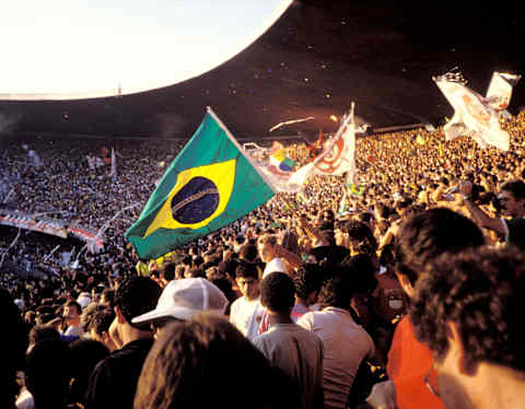 A large crowd fills a stadium, many waving flags, including a prominent Brazilian flag in the centre. The stands are packed with cheering fans, and the atmosphere appears festive and lively.