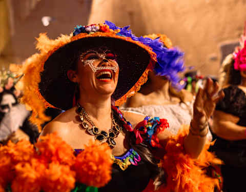 A woman in a flamboyant skirt and hat in pinks and purples, with marigold-orange feathers, enjoys the Dia de Muertos parade.