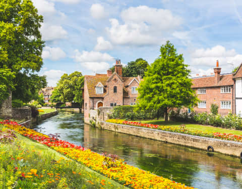 The River Stour flows past the historic, half-timbered houses of Canterbury, bordered by lawn banks of red and gold flowers.