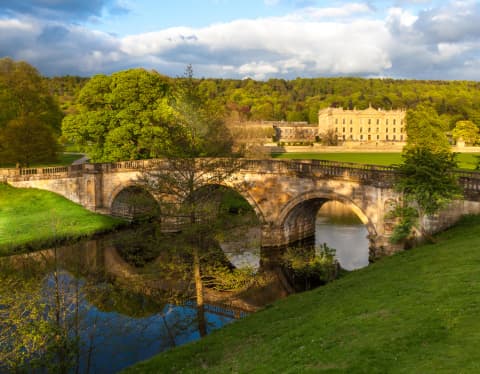 A three-arch stone bridge bows overs the River Derwent as it flows through lush parklands, overlooked by Chatsworth House.