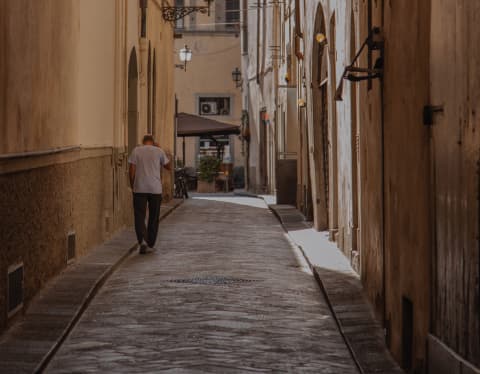 A man walks along a narrow cobbled back street in the shade of tall buildings in an old Italian city.