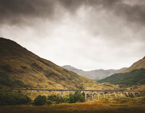 With the Scottish Highland's mountains looming, the arches of the Glenfinnan Viaduct span a moss and heather-green valley.