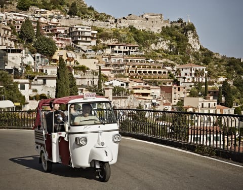 A classic white Piaggeo Ape offers guests great views beyond its open red cabriolet roof, as it explores Taormina's streets