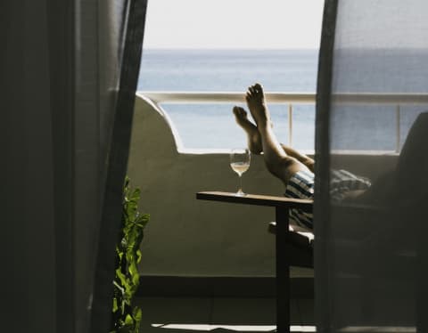 Partly obscured by diaphanous drapes, a man reclines in a chair on his sea-view balcony with a glass of wine and his feet up.