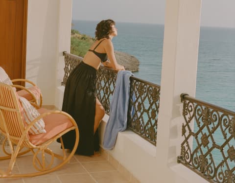 On a balcony with wicker chairs, a woman in a black bikini top and skirt leans on the filigree railings, gazing at the sea.