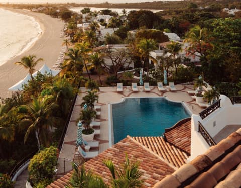 Sunset glows on the sand, roof tiles, palm trees and pool in a birds-eye view of the resort and distant sweep of Baie Longue.