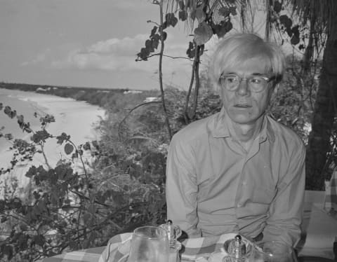 In a black and white image taken during his stay, artist Andy Warhol sits at a cliff-top table with views over the beach.