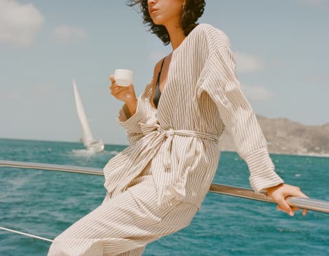 A woman enjoys an onboard espresso during a day at sea, as a keeling sailboat behind glides through the aquamarine seascape.