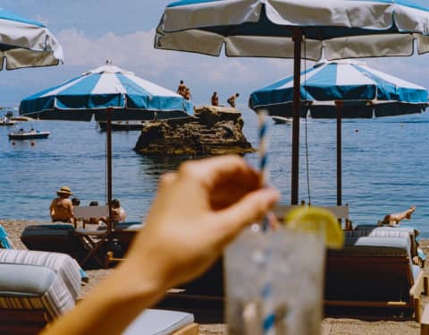 A guest stirs a lemon-garnished drink in soft-focus foreground as swimmers gather on a rock out to sea, beyond the parasols.