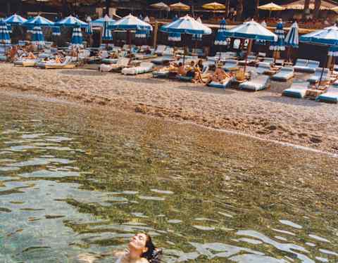 In front of the beachfront parasols, two women in  swimming costumes sit in the shallows, heads tipped back, seen from above.