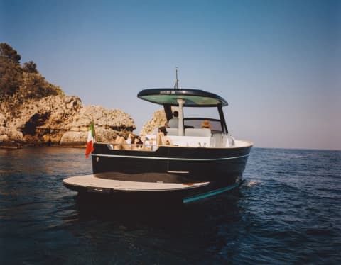 A couple sunbathes on the lounge deck of an Apreamare Gozzo boat as sails in full sun towards the headland, seen from behind.