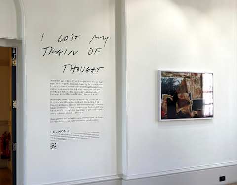 A white gallery wall displays text reading “I lost my train of thought” above a block of smaller print. To the right, a framed artwork hangs above a wooden floor, under ceiling track lighting.