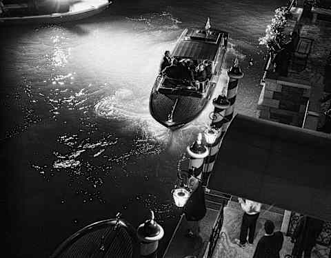 In darkness, hotel staff wait as a late boat pulls up alongside the striped paline posts, guided by lamps, seen from above.