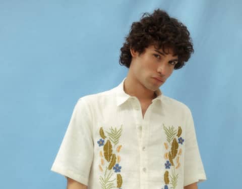 A model poses with an orange in blue trousers and an off-white linen shirt with floral embroidering by  Amor & Rosas.