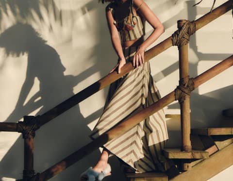 A model in a cream and brown striped skirt and crop-top by Sandra Weil descends wooden stairs dappled by fern shadows.