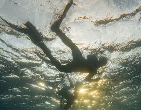 Looking up a the silhouettes of two snorkelers swimming with flippers through surface sun rays, viewed from depths below.