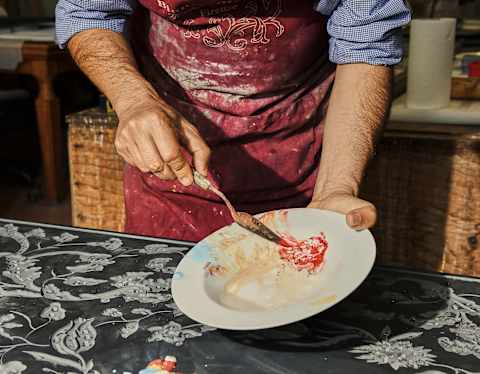 A person in a red apron stands at a table covered with a black and white floral cloth, mixing paints on a white plate using a brush. The person's face is not visible.