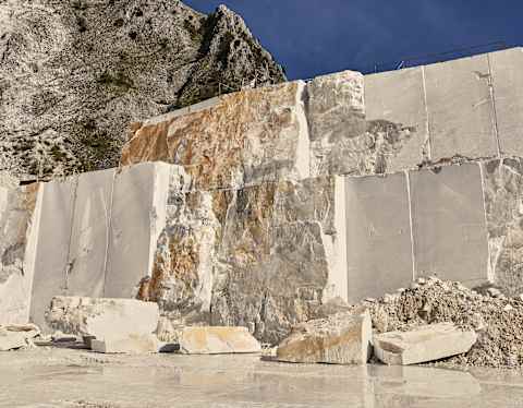 Large marble blocks are cut from a quarry wall, with rough and smooth surfaces visible. The quarry is set against rocky hills under a clear blue sky, and loose stone debris covers the ground.