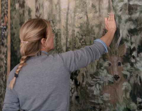 A person with blonde hair in a plait is facing away, painting or restoring a large, detailed mural or tapestry depicting a forest scene with tall trees and dense foliage.