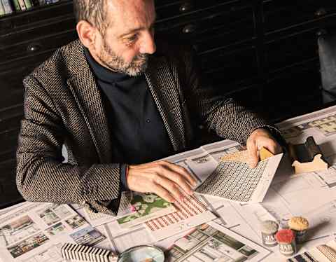 A man sits at a desk covered with architectural drawings, a magnifying glass, pens, and building models, examining a sample tile in his hands.