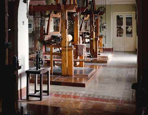 A view through an arched doorway reveals a room with large wooden weaving looms, natural light filtering in, and tiled floors, suggesting a traditional textile workshop.