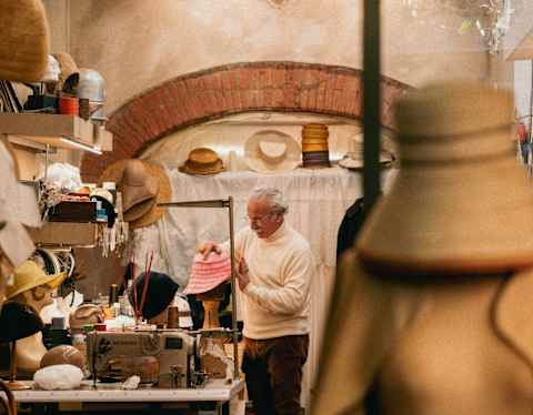 A man stands in a cosy workshop filled with hats and sewing materials. He appears to be working or contemplating, surrounded by shelves with hats and a sewing machine. The foreground shows a hat and coat on display.