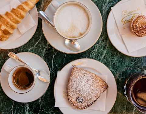 An overhead view of a marble table set with coffee, a cappuccino, a croissant, and two pastries dusted with icing sugar on white plates, with serviettes and a packet labelled “Gualtieri”.