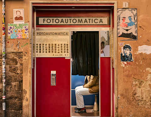 A person sits inside a red photo booth with a black curtain, set in an old, orange wall decorated with graffiti, posters, and a white angel illustration above the booth labelled Fotoautomatica.