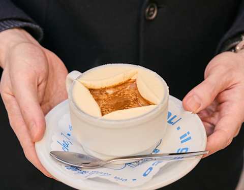 A person holds a white cup of coffee or cappuccino on a matching saucer with a teaspoon. The coffee has a square-patterned froth on top. The person is wearing a dark coat.