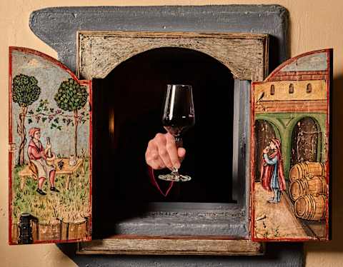 A hand holding a glass of red wine reaches through a small wooden window with painted doors depicting historical scenes, set in a stone frame on a beige wall.