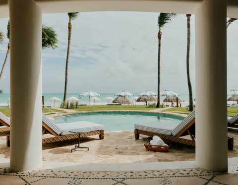 Four loungers surround a private pool set back from the sand, sea and beach parasols, seen from Villa Maroma's colonnade.