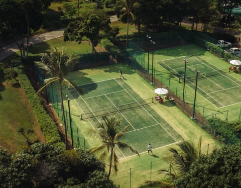 An aerial shot of a man playing tennis on one of two dark green courts with lime grass surrounds and white parasol shades.