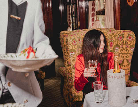 A young woman in a red jacket sits at a table on a vintage train, holding a glass, whilst a waiter in a white uniform brings pudding. The decor is ornate, with patterned chairs and wood panelling.