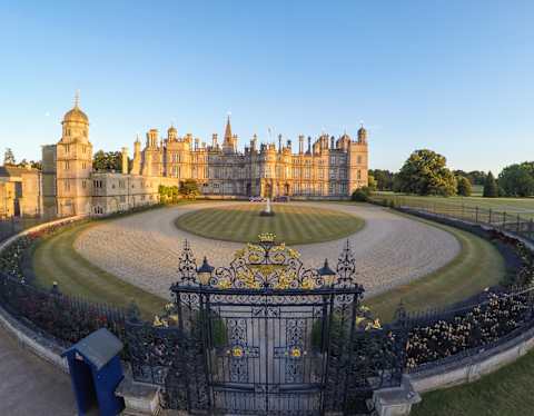 High-angle view of the large Burleigh House estate, stone glowing gold in late sun, seen over the wrought iron gates.