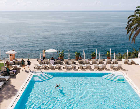 Two guests swim in the large, sparkling blue pool, which offers uninterrupted views of deeper ocean hues, seen from above.